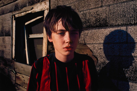 A color portrait photograph of a White boy with brown hair wearing a red and black striped shirt, standing in front of a building partially covered in roof shingles.