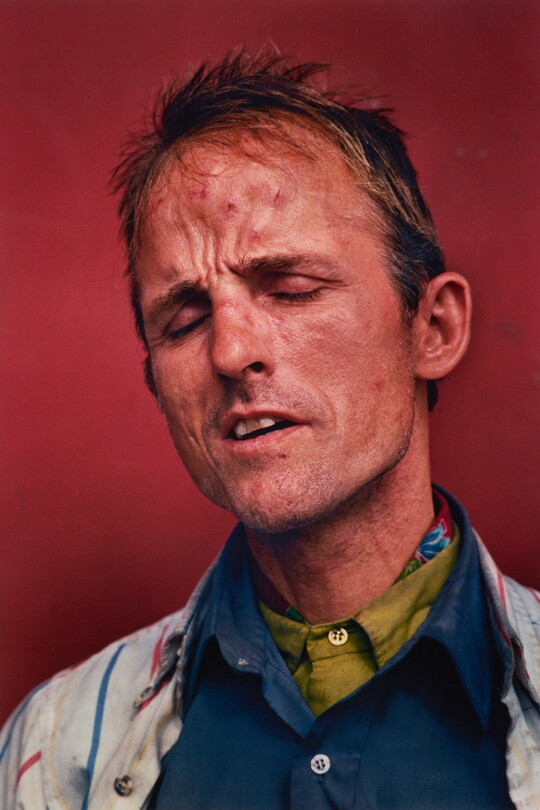 A color portrait photograph of a White man with his eyes closed and mouth slightly open wearing several layers of collared shirts.