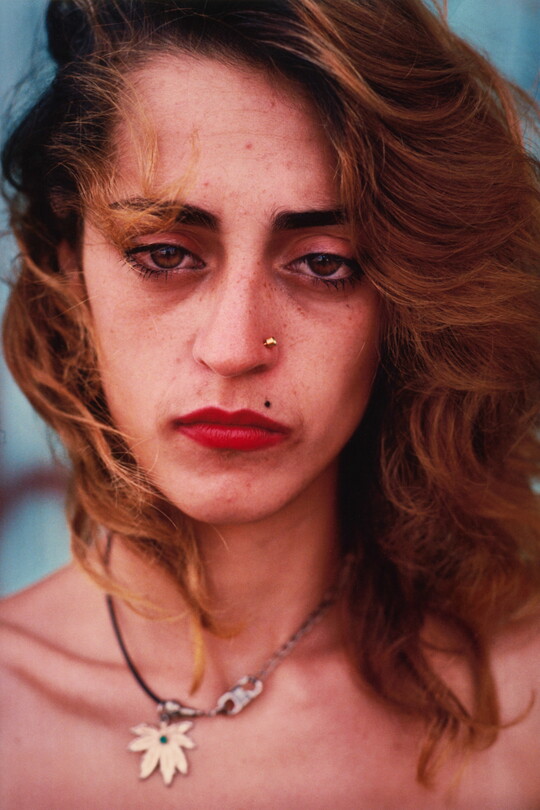 A color portrait photograph of a White woman with brown eyes, wavy shoulder-length brown hair, a gold stud in one nostril, red lips, and wearing a hemp leaf necklace.
