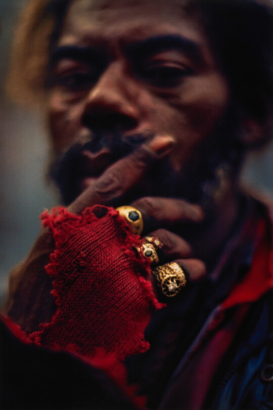 A color portrait photograph of a Black man with a mustache, face slightly out of focus, resting his chin in one hand; on his hand are three large gold rings and a torn red glove.