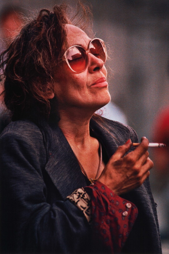 A color portrait  photograph of a medium-skinned woman with short brown hair wearing large sunglasses, a navy blue blazer, and holding a cigarette.