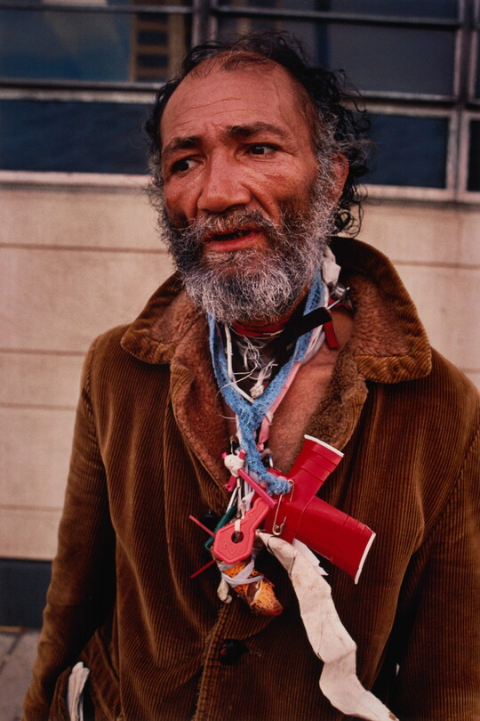 A color portrait photograph of a balding Black man with graying beard wearing a brown coat and random objects around his neck.