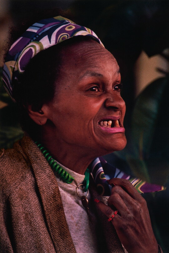 A color portrait photograph of a Black woman, smiling so that her underbite and missing teeth show, wearing a colorful scarf in her hair.