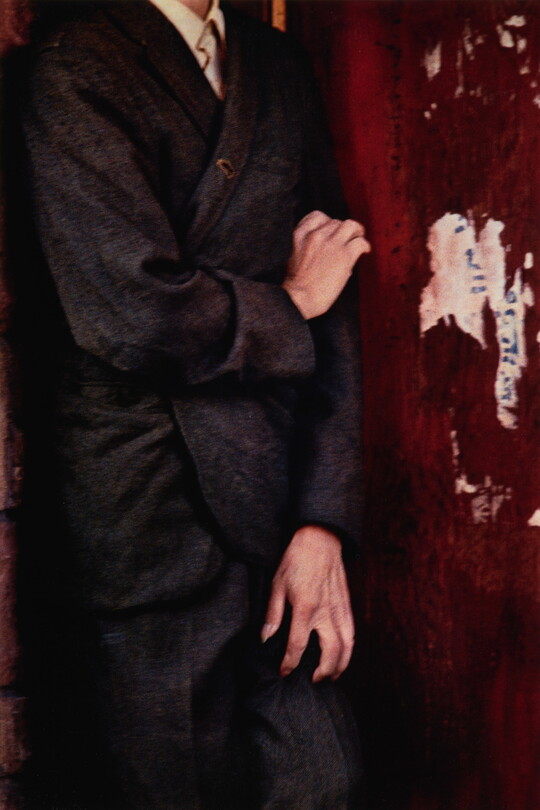 A color photograph of a White person from the neck down, wearing a loose-fitting suit, clawing at their leg and arm with their hands.