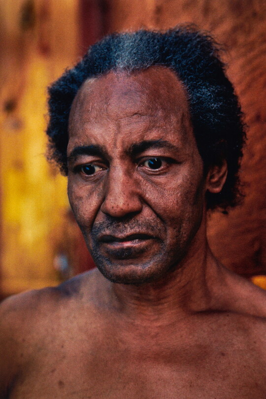 A color portrait photograph of a Black man with salt and pepper hair, stubble on his face, a crease in his forehead as if worried, looking downward.