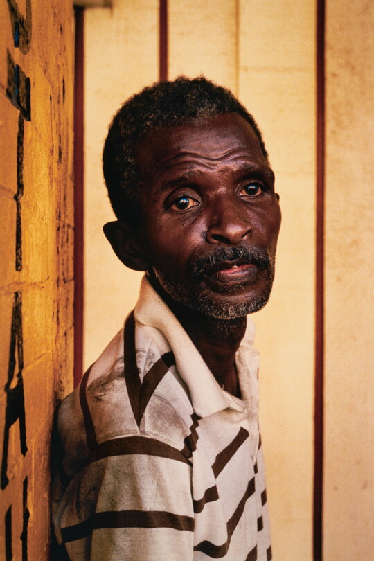 A color portrait photograph of an older Black man, short hair and stubble on his thin face, wearing a striped polo shirt, and leaning against a wall.