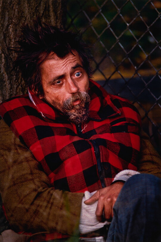 A color portrait photograph of a White man, seated against a tree, with messy dark hair, misshapen eyes and a graying goatee, wearing a red-and-black checked vest.