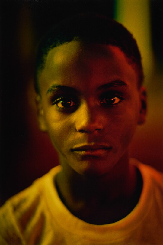 A color portrait photograph portrait of a young Black person, close-cropped hair, wearing a white t-shirt; yellow lighting casts tinted shadows across their face.