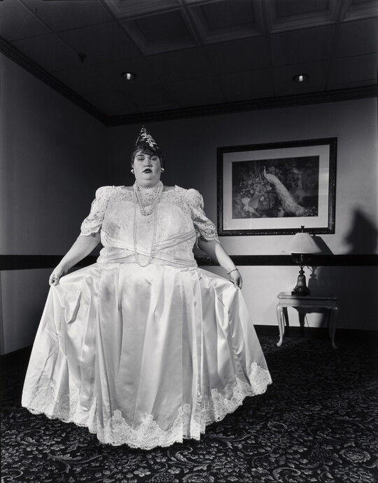 A black-and-white photograph of a White woman holding out the skirt of her long, formal white dress.
