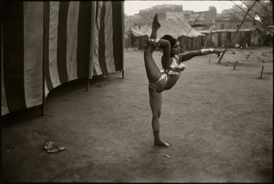 A black-and-white photograph of a dark-skinned woman with dark hair in a sequined bikini and metallic cuffs stretching her leg behind her head as she stands next to a striped tent.