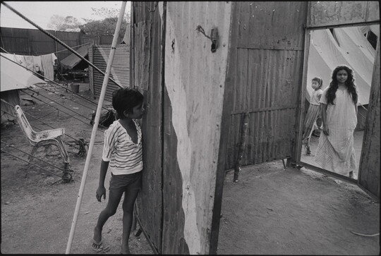A black-and-white photograph of a dark-skinned boy peeking through a hole in a wooden structure as two dark-skinned girls appear in a doorway on the opposite side.