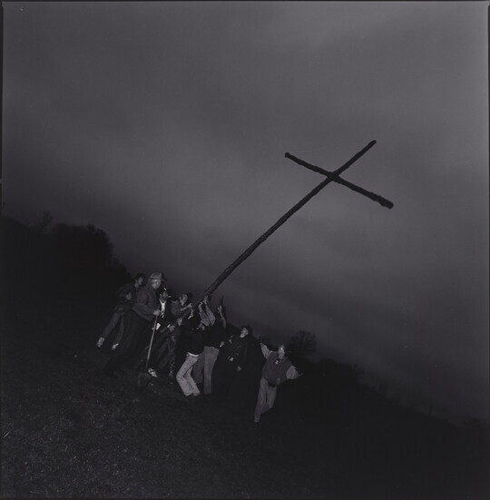 A black-and-white photograph of a group of people hoisting up a large cross in a field at dusk.