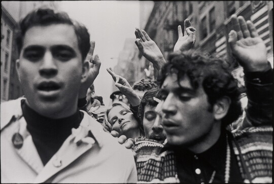 A black-and-white photograph of two people amongst a crowd of protesters, some of whom are holding up their hands and making circles with their index fingers and thumbs.