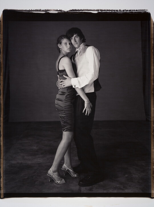A black-and-white photograph of a young White couple in formal attire embracing one another and looking at the camera.