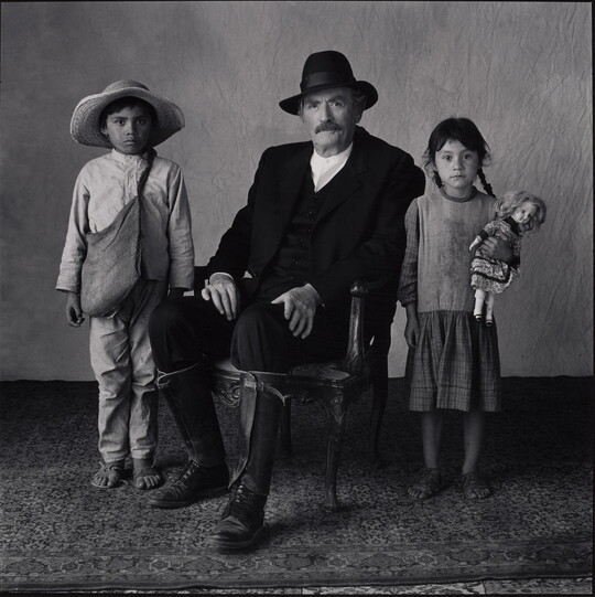 A black-and-white portrait photograph of a seated White man with a moustache, wearing a hat, black suit, and tall boots with two medium-skinned children standing on either side of him.