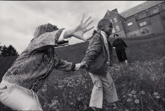 A black-and-white photograph of two children holding hands and playing in a grassy field with a Black woman and large brick building in the background.