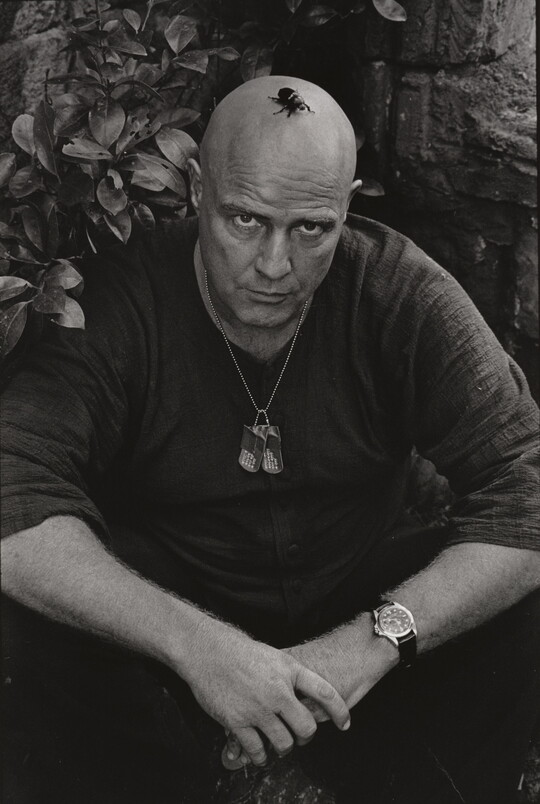 A black-and-white photograph of a bald White man seated on the ground with his arms resting on his knees and a large beetle on his head.