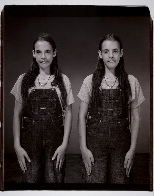 A black-and-white photograph of White twin girls with long dark hair, both in overalls standing side-by-side in the same pose.