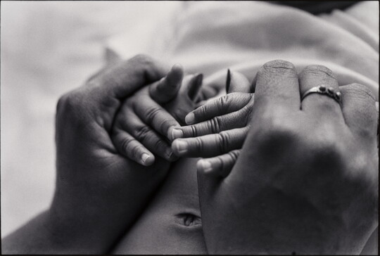 A close-up, black-and-white photograph of adult Black hands clasping Black baby hands.