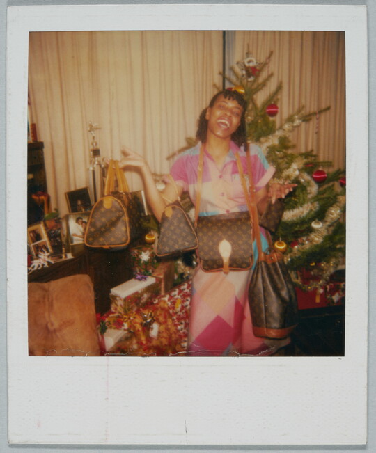 A color Polaroid photograph of a Black woman gleefully showing off several designer purses in front of a Christmas tree.