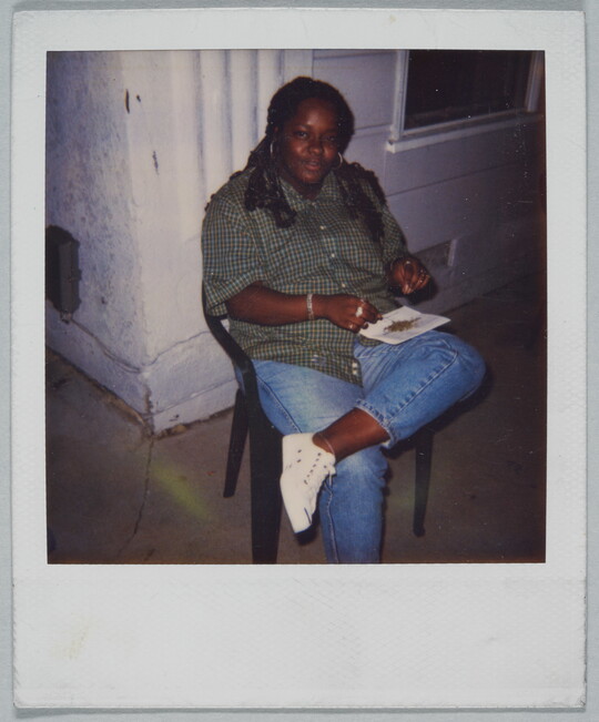 A color Polaroid photograph of a Black woman seated on a plastic chair balancing a piece of paper with bits of something on it on her crossed leg.