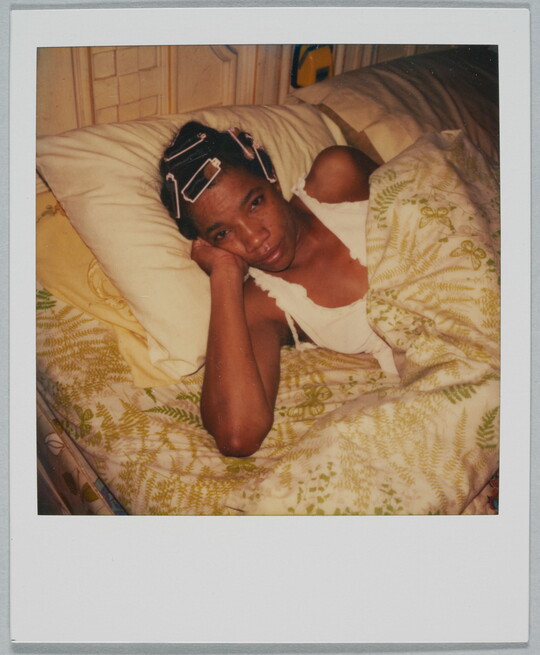 A color Polaroid photograph of Black woman, curlers in her hair, in bed with her head propped up on her hand.
