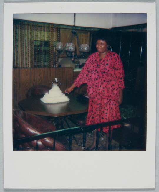 A color Polaroid photograph of a smiling Black woman in a pink and black dress slicing a tiered white cake on a dining table.