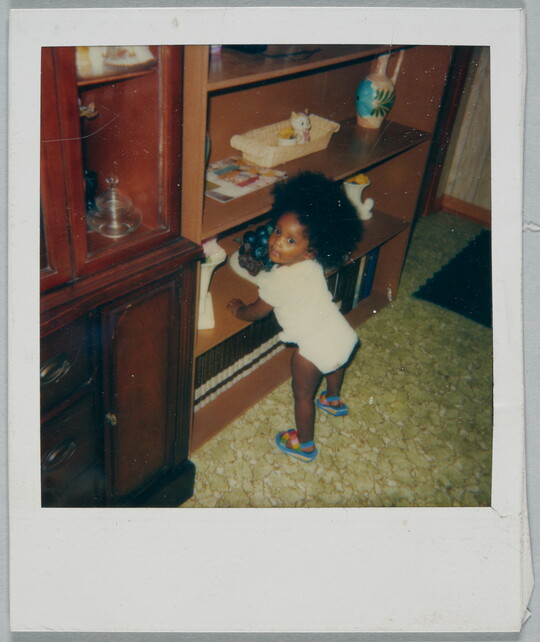 A color Polaroid photograph of Black toddler in a shirt, diaper, and sandals with big, natural hair propping themselves up on a bookshelf.