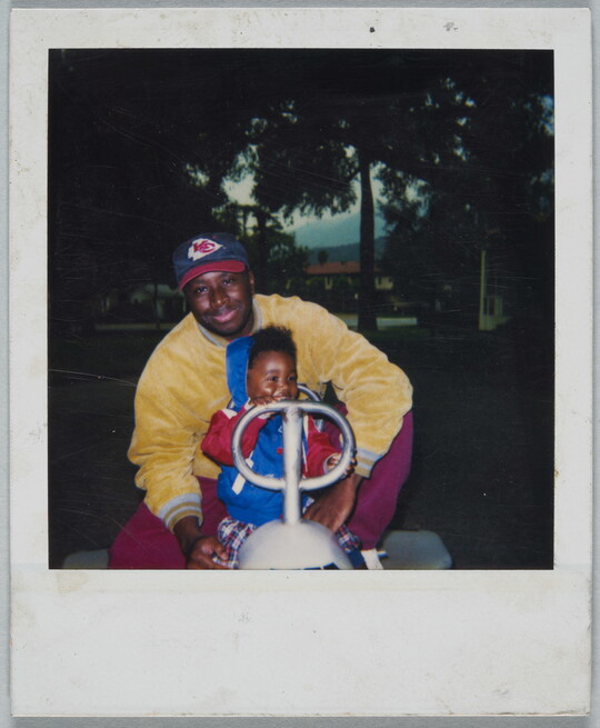 A color Polaroid photograph of a Black man and toddler seated behind the handles of an outdoor play toy in a park.