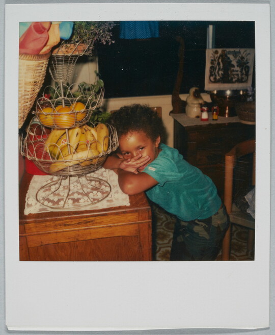 A color Polaroid photograph of a Black child leaning on a table that has a tiered fruit holder.