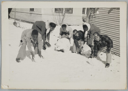 A black-and-white snapshot of smiling Black children building snowmen next to a house.