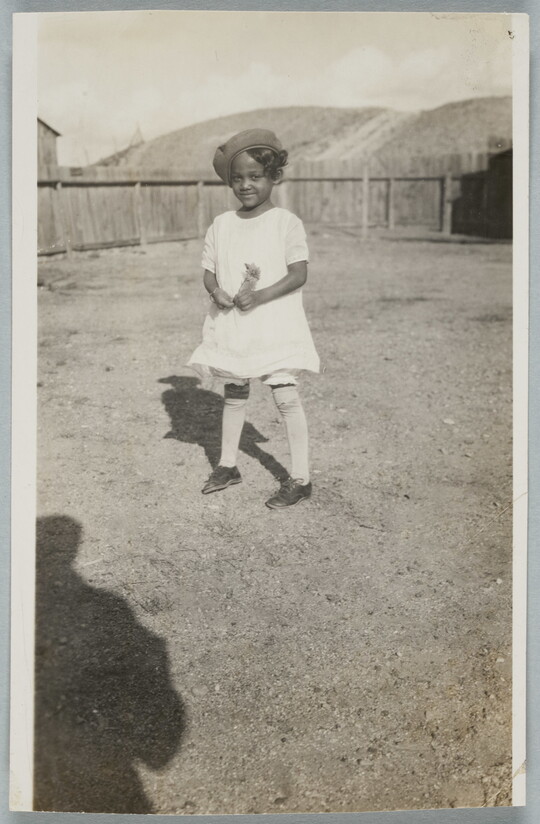 A black-and-white snapshot of a Black girl in a white dress and knee socks standing in a fenced yard.