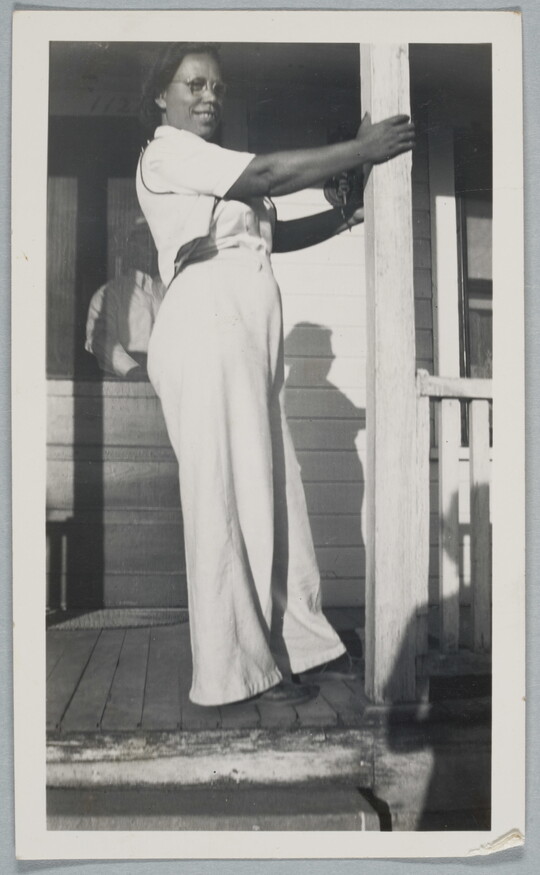 A black-and-white snapshot of a Black woman in white shirt and pants standing on a porch holding onto a column.