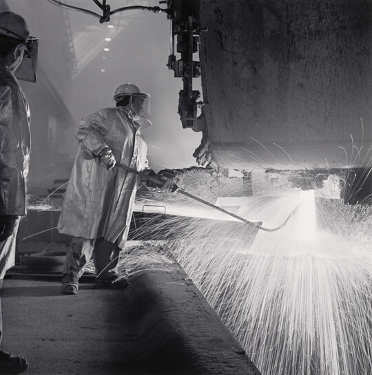 A black-and-white photograph of an industrial worker, wearing safety helmet, coat, and gloves holding a rod under a machine emitting sparks.