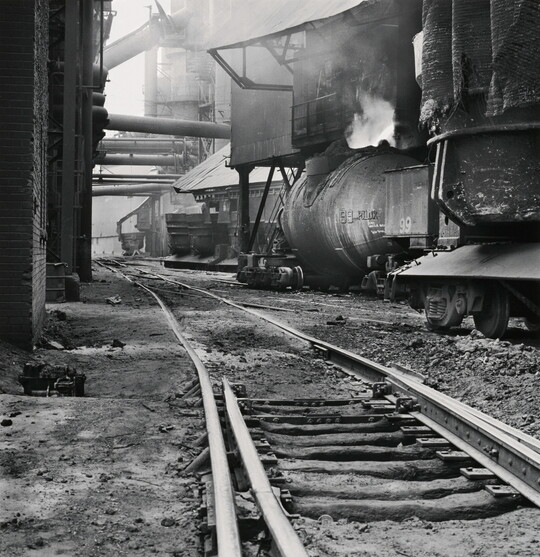 A black-and-white photograph of railroad track  between industrial buildings and  machinery, some emitting steam or smoke.