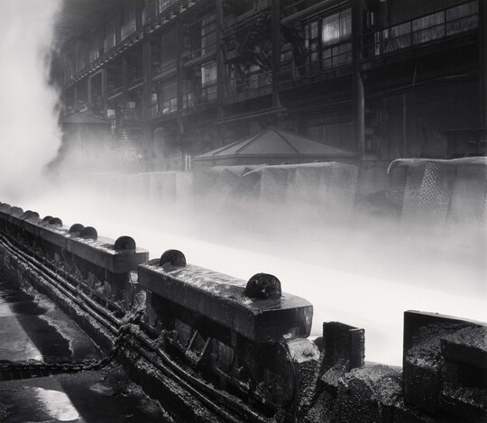 A black-and-white photograph of industrial machinery in front of steam or smoke rising near a multi-story industrial building.