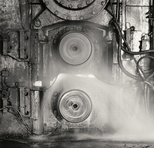 A black-and-white photograph of two spinning metal discs set into a metal wall that has pipes and tubes connected to it.