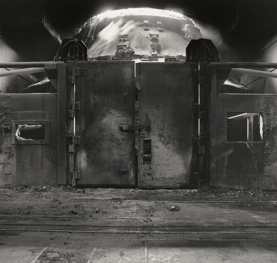 A black-and-white photograph of a metal wall with large doors behind which is an industrial furnace.