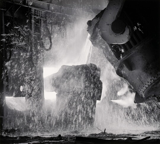 A black-and-white photograph of a piece of machinery pouring molten metal into another piece of machinery, all surrounded by sparks.