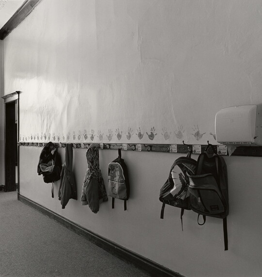 A black-and-white photograph of a hallway with kids' backpacks and jackets hanging on hooks.