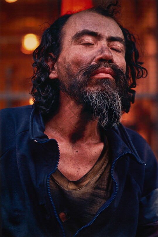 A color portrait photograph of a medium-skinned man with black hair and goatee, eyes closed, dirt smudged on his face, wearing a dark blue jacket.