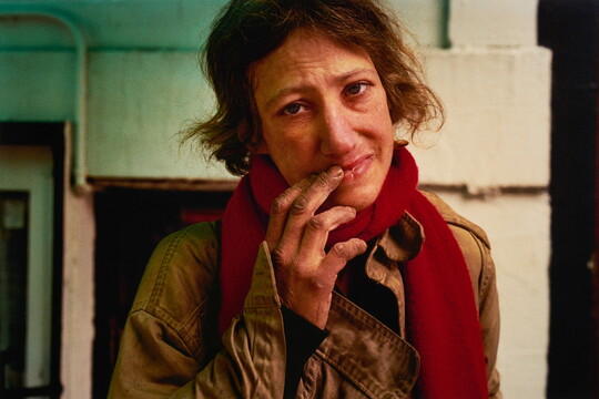 A color portrait photograph of a White woman with short brown hair, wearing a red scarf over a green jacket; one of her hands is raised to touch her lips and her expression is tired and weary.
