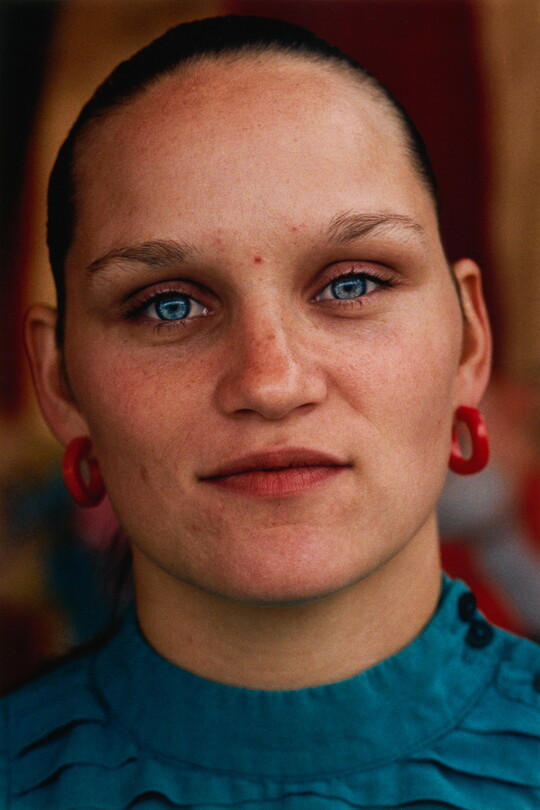 A color portrait photograph of a White person with light blue eyes, hair pulled back tightly on their head, wearing red hoop earrings, and a teal top.