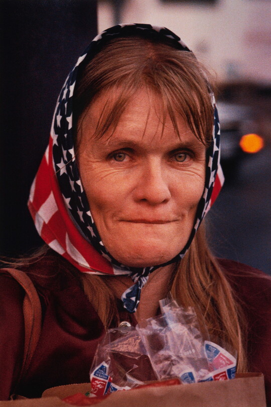 A color portrait photograph of a middle-aged White woman with shoulder-length hair, prominent cheekbones and creases around her eyes, wearing an American flag scarf around her head.