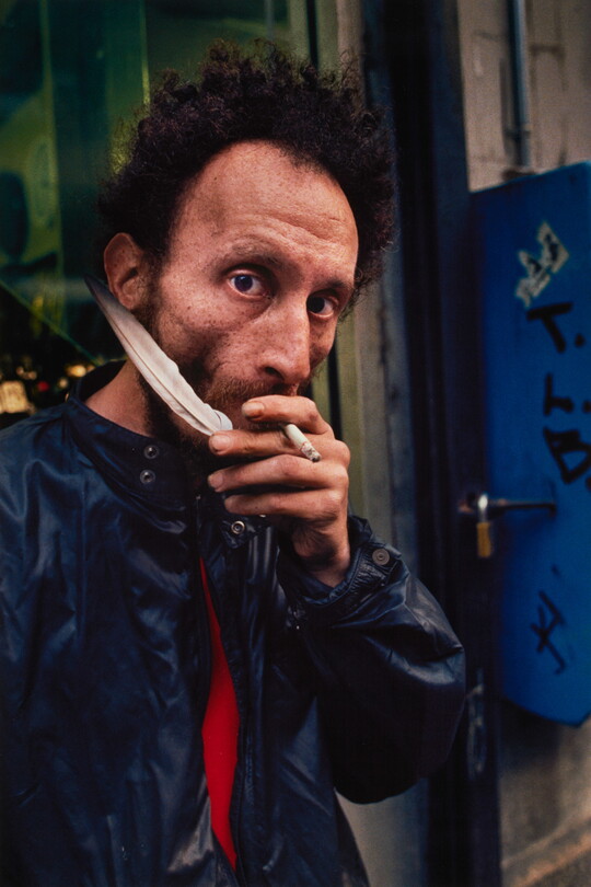 A color portrait photograph of a medium-skinned man with curly hair, holding a cigarette in his mouth and a white feather in his hand, wearing a blue jacket.