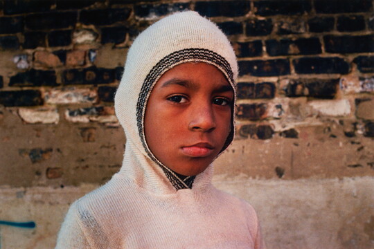 A color portrait photograph of a young dark-skinned child wearing a white hooded sweater with the hood pulled tight around their head.