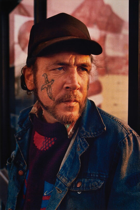 A color portrait photograph of a middle-aged White man with a beard and mustache and a sword tattoo on his cheek, wearing a dark baseball cap and jean jacket.