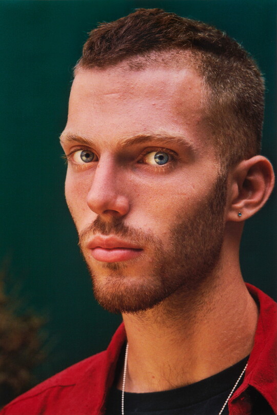 A color portrait photograph of a young White man with blue eyes, very short hair, beard and mustache, wearing a stud in one ear and a black shirt under a red button-down shirt.