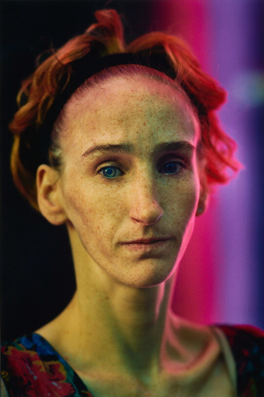 A color portrait photograph of a White woman with a headband pulling back her short auburn hair, a freckled face, and prominent cheekbones.