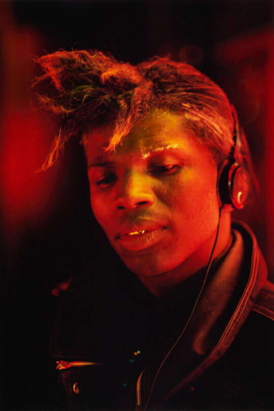 A color portrait photograph of a Black man, hair pulled into a sort of ponytail over his forehead, wearing headphones, bathed in reddish-orange light.